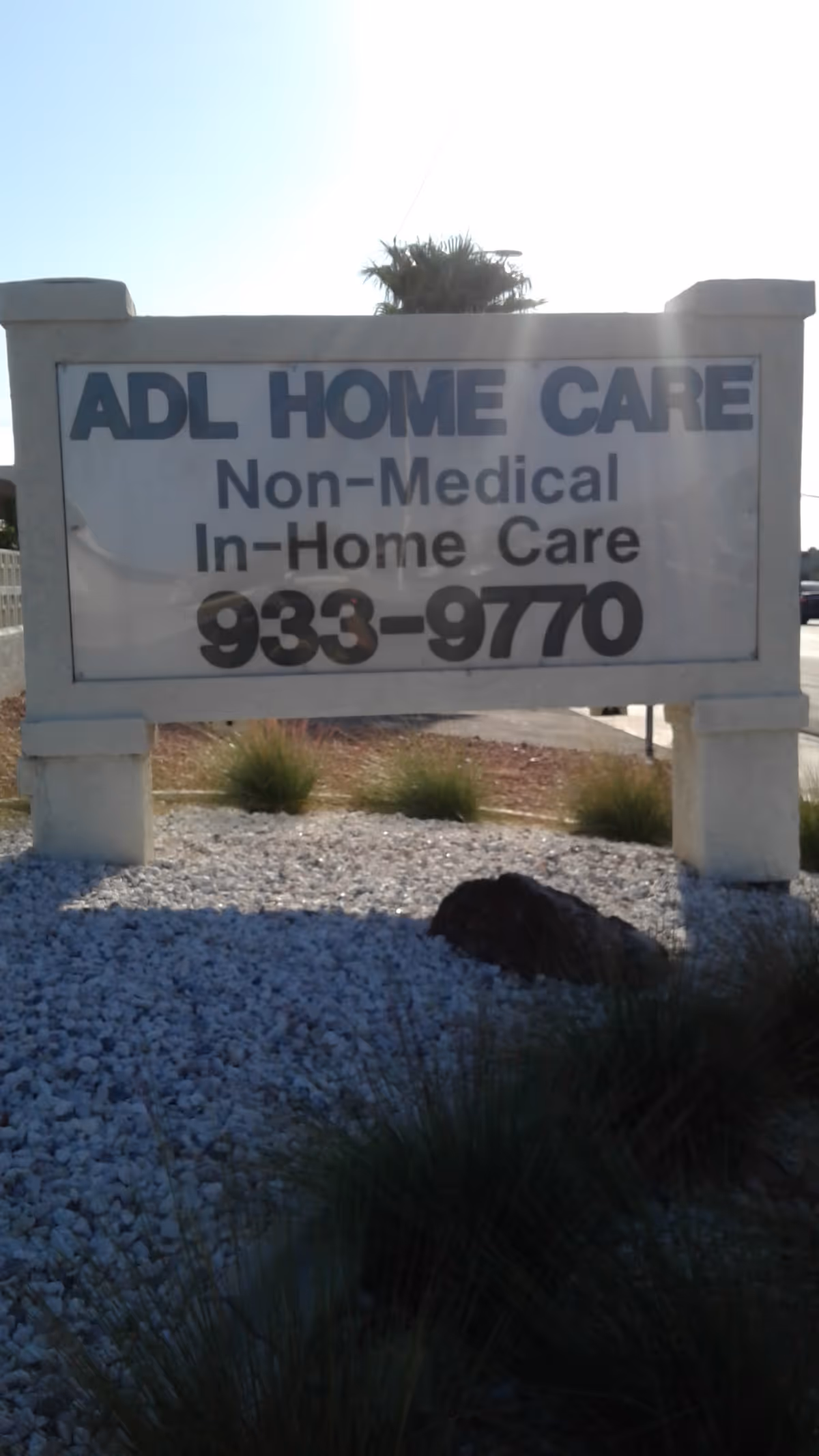 Outdoor sign advertising ADL Home Care with contact number, set among rocks and shrubs.