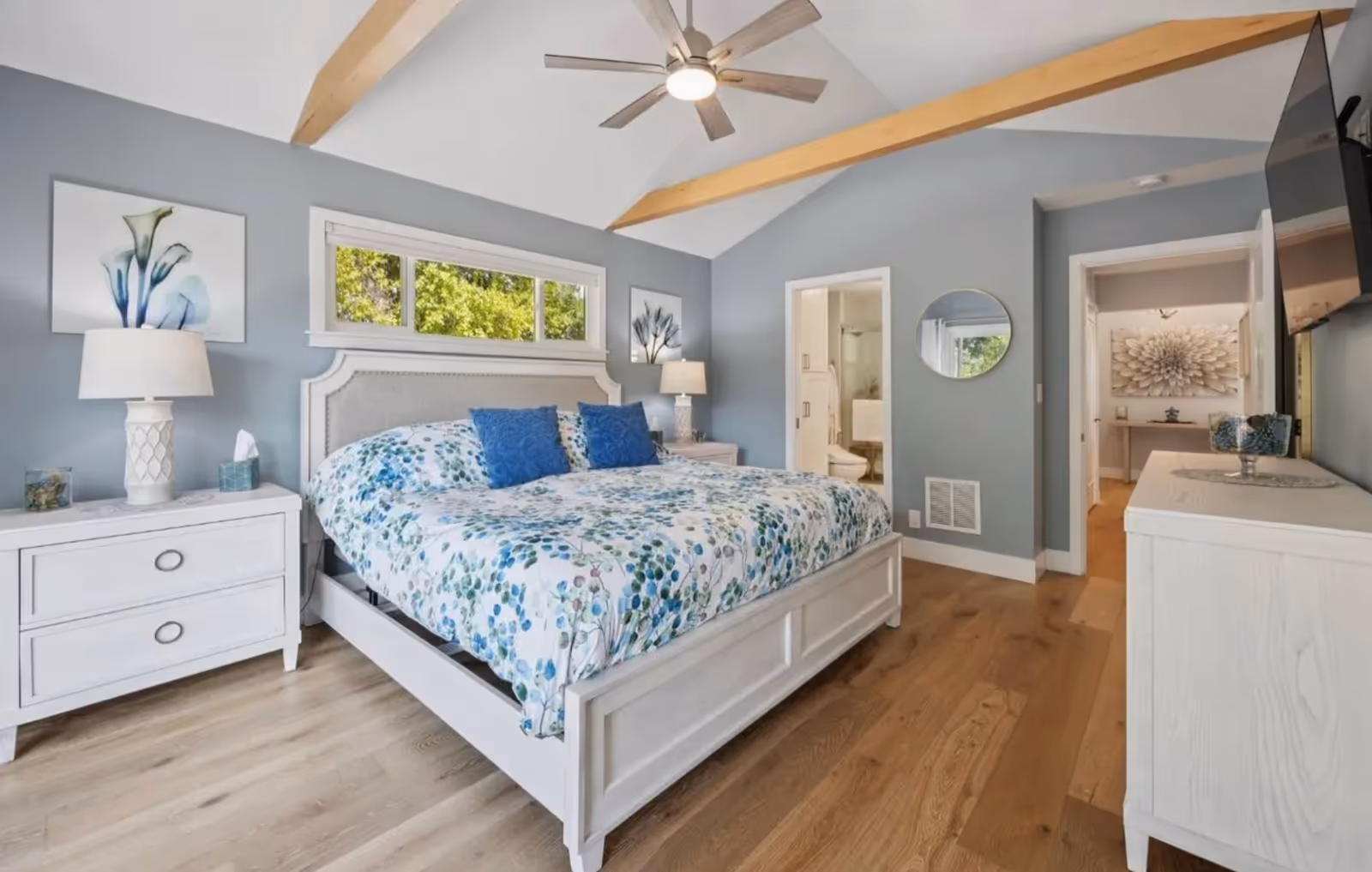 Bright bedroom with white furniture, blue walls, ceiling beams, and floral bedding.
