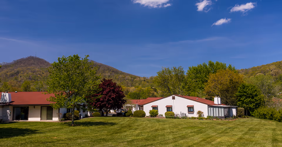 Single-story white retirement community buildings with red roofs on a large mowed lawn, trees, and hills in the background.
