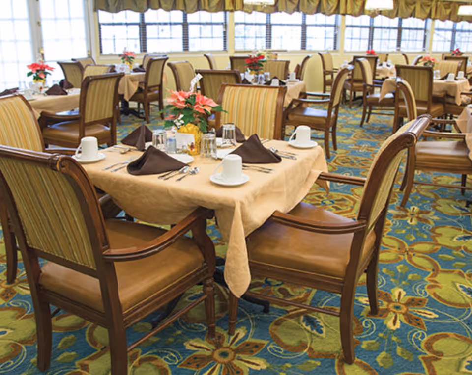 Bright dining room with multiple tables set with beige tablecloths, brown napkins, wooden chairs, and floral centerpieces.