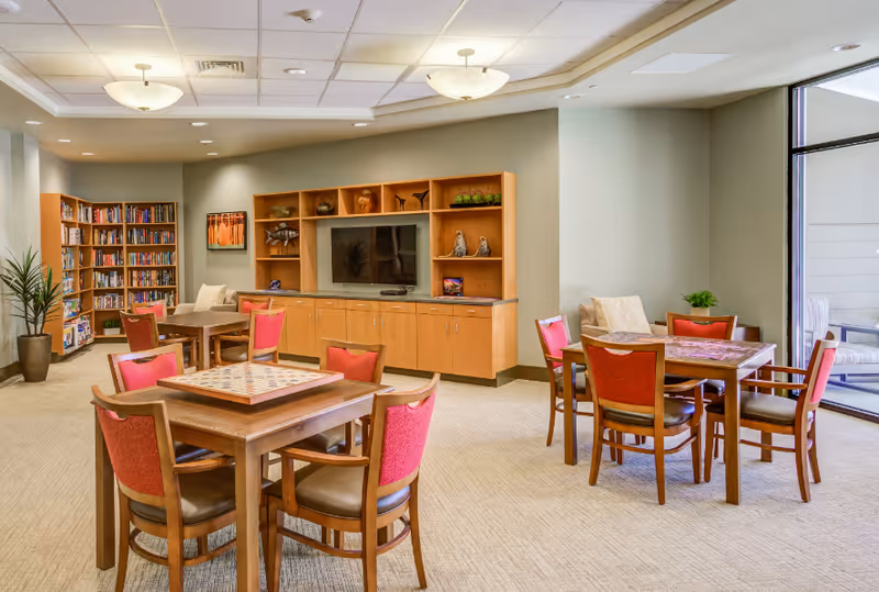 Bright senior living lounge with tables, red chairs, a TV wall, bookshelves, and large windows.