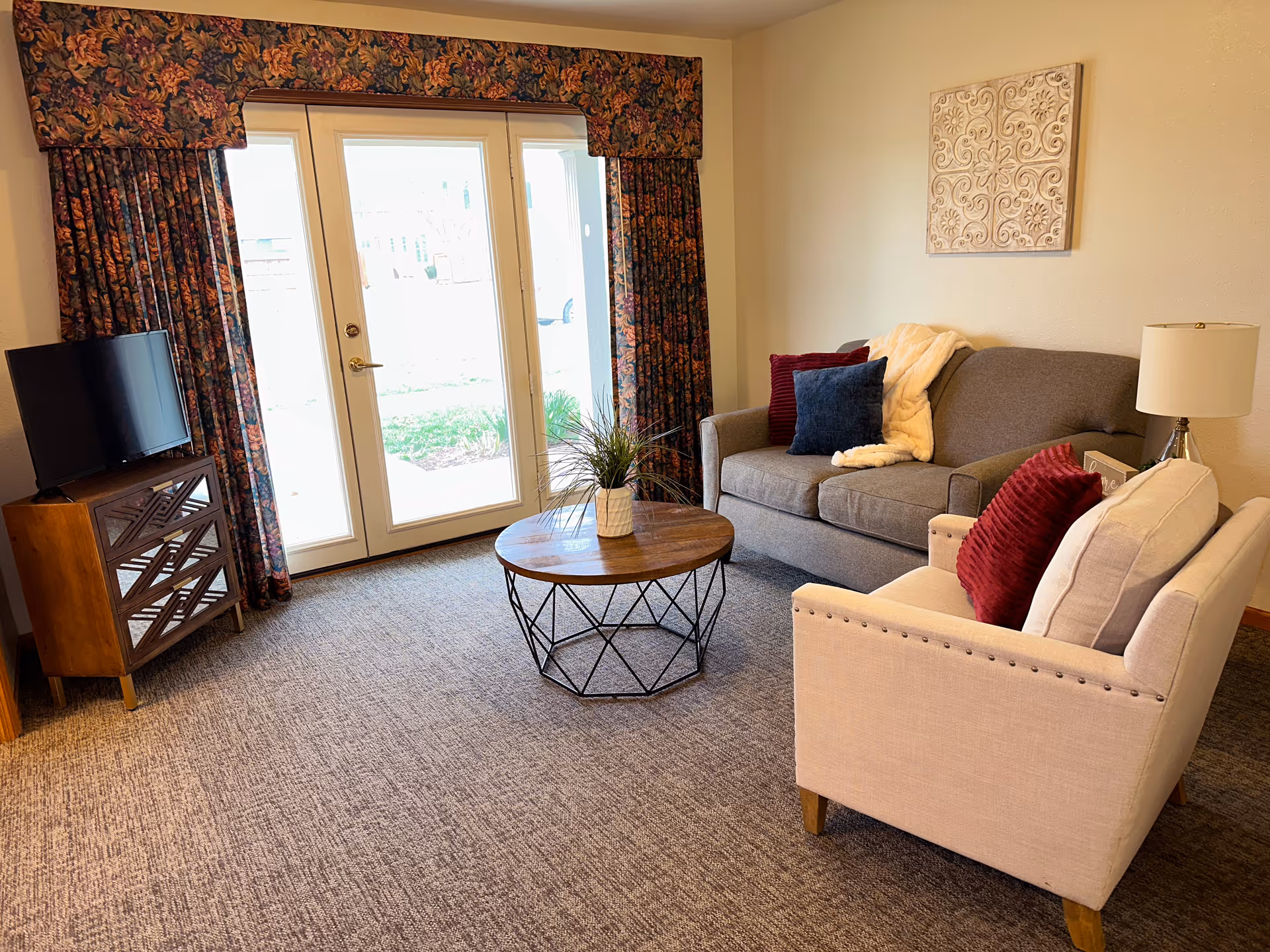 A cozy living room with a gray sofa adorned with red and blue pillows and a cream-colored throw blanket. A beige armchair with a red pillow sits nearby. A round wooden coffee table with a metal frame holds a small potted plant. A wooden TV stand with a flat-screen TV is positioned near glass double doors covered with floral-patterned curtains. A table lamp and decorative wall art complete the room.