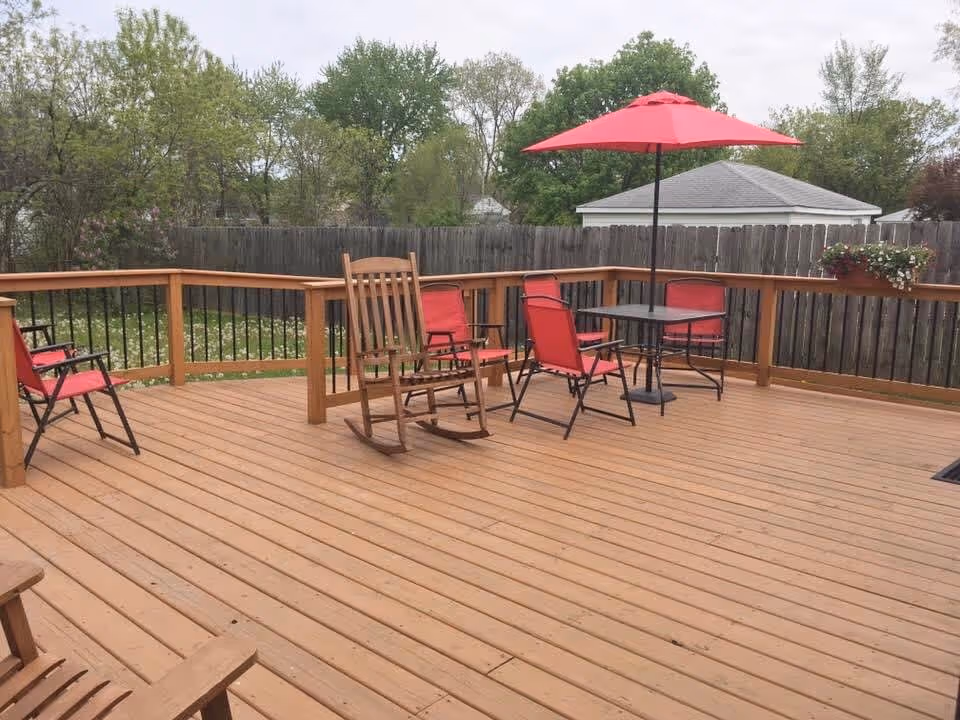 Outdoor wooden deck area with several red cushioned chairs, a wooden rocking chair, a small black table with a red umbrella, and a wooden fence surrounding the yard with trees and a small white shed in the background.