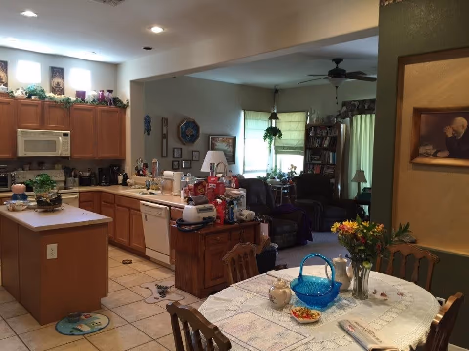 View of a cozy kitchen and dining area in a senior living facility. The kitchen features wooden cabinets, a microwave, dishwasher, and various kitchen items on the counters. The dining area has a round table covered with a lace tablecloth, decorated with a blue basket, a vase of flowers, and other small items. In the background, a living room area with armchairs, a bookshelf, and windows with blinds is visible.