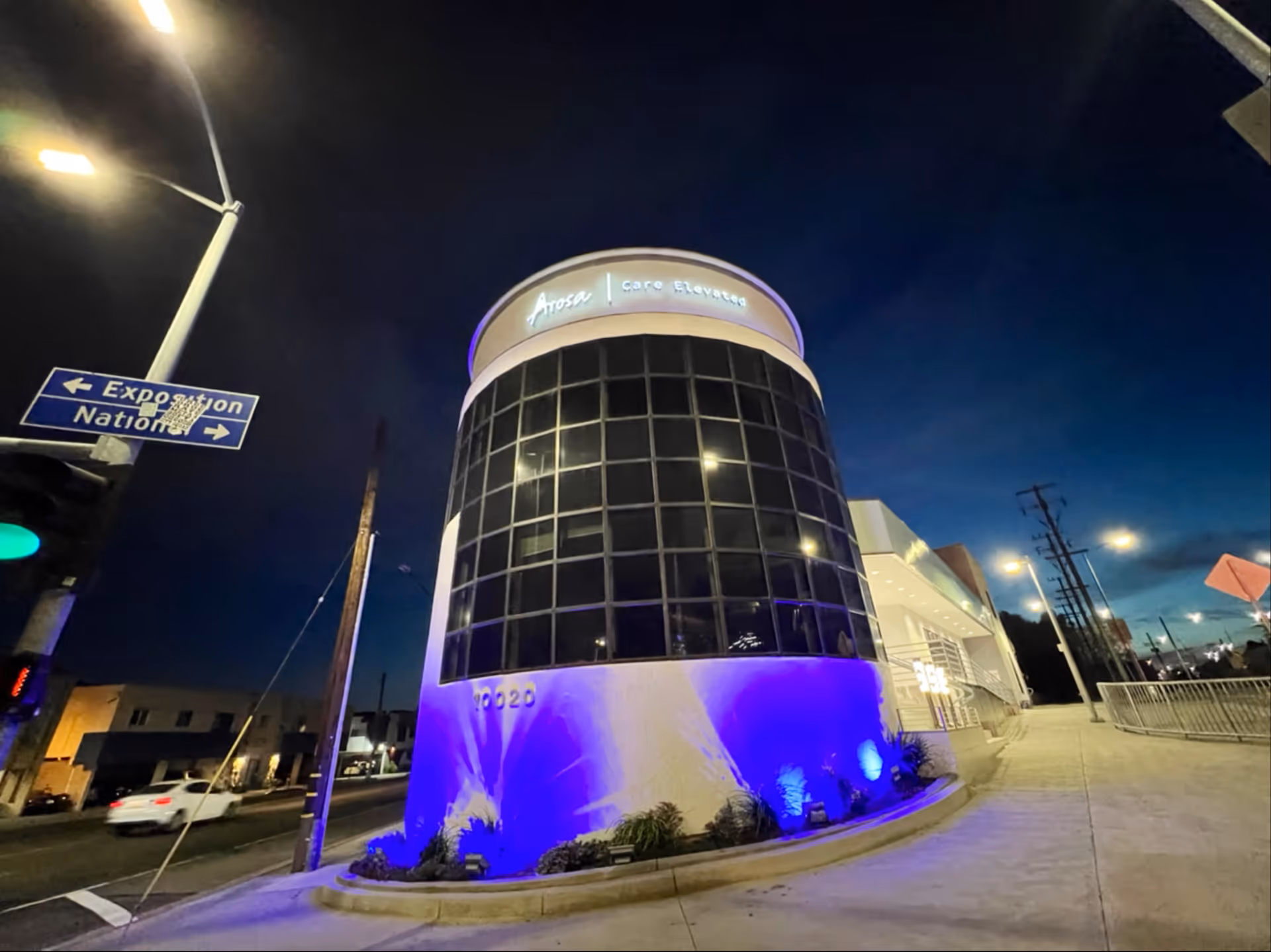 Night exterior of Arosa Los Angeles facility with curved glass facade and blue lighting