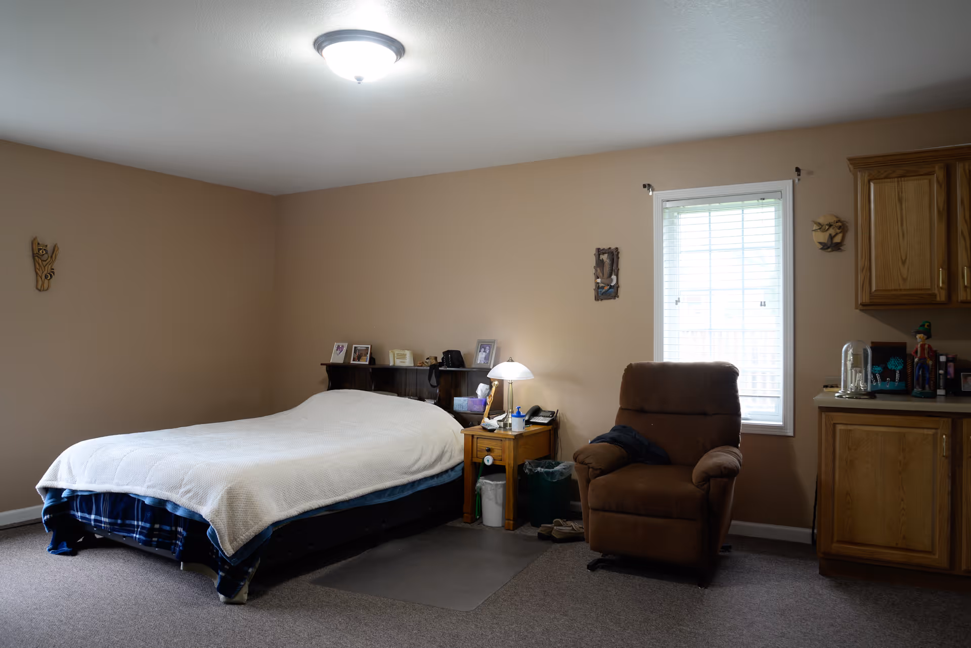 Bedroom with a made bed, wooden nightstand and lamp, a brown recliner by a window, and oak cabinets.