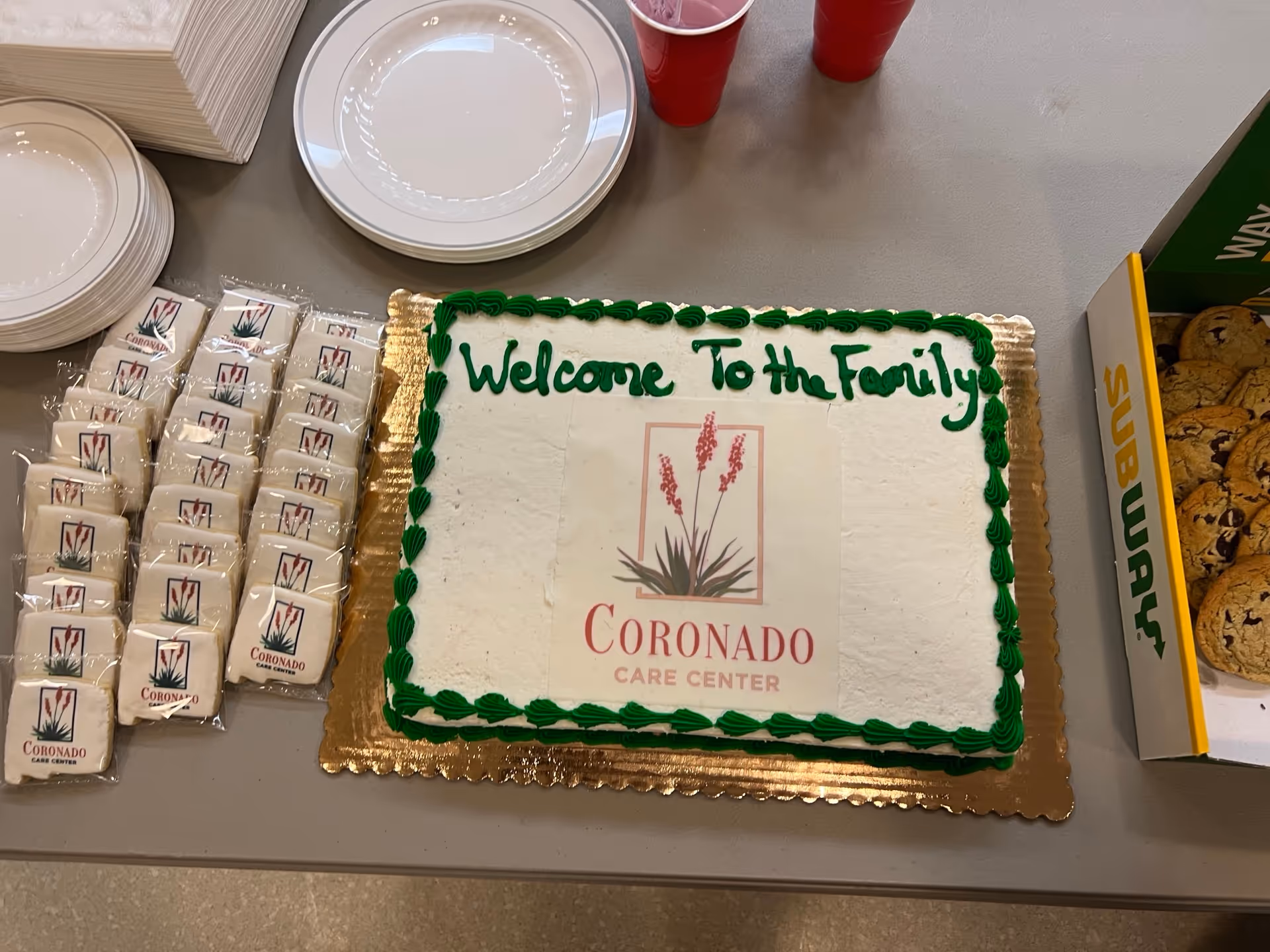 Cake reads 'Welcome To The Family' beside Coronado Care Center logo on a serving table.