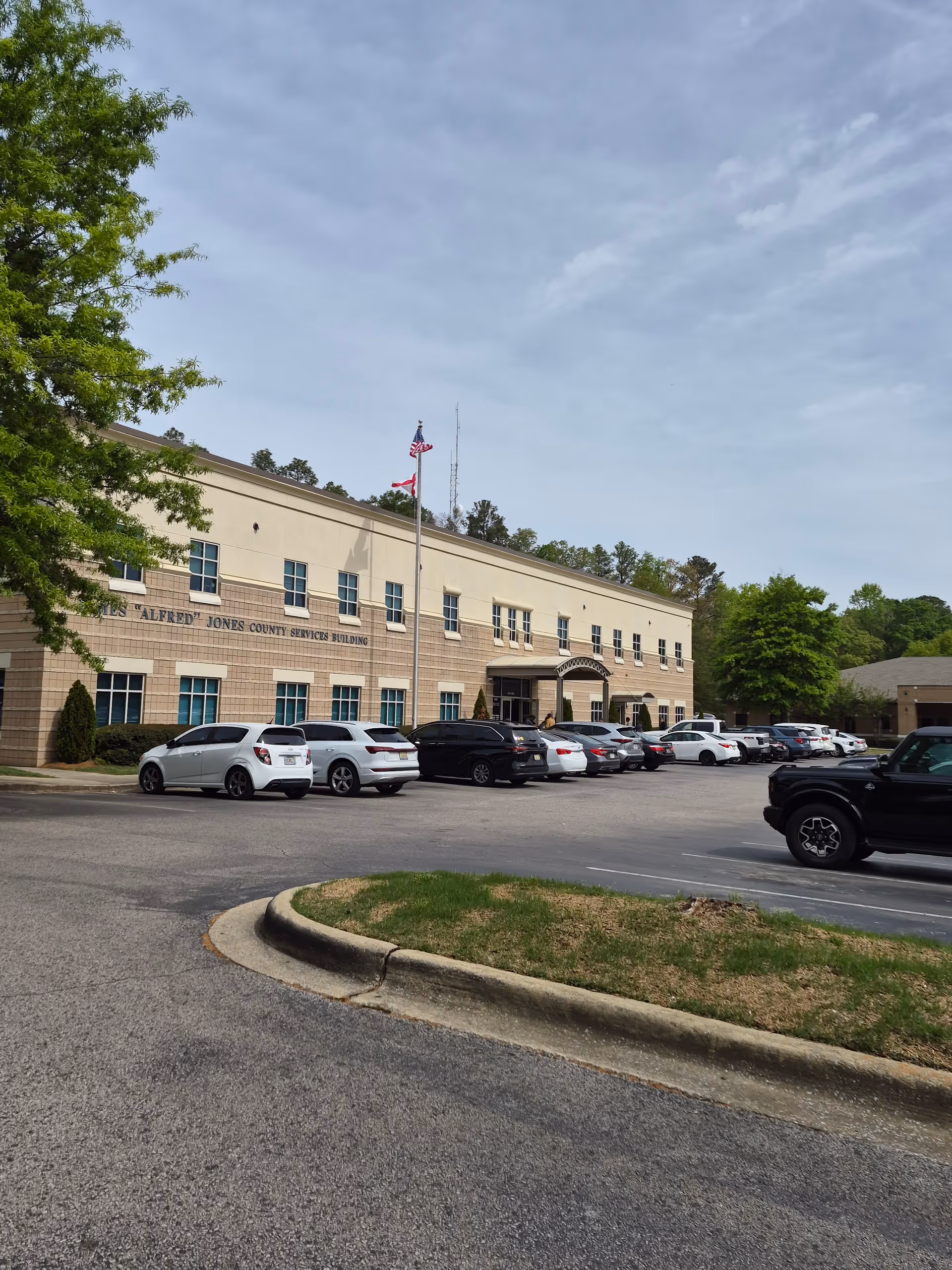 Two-story beige government building with a parking lot and several parked cars.