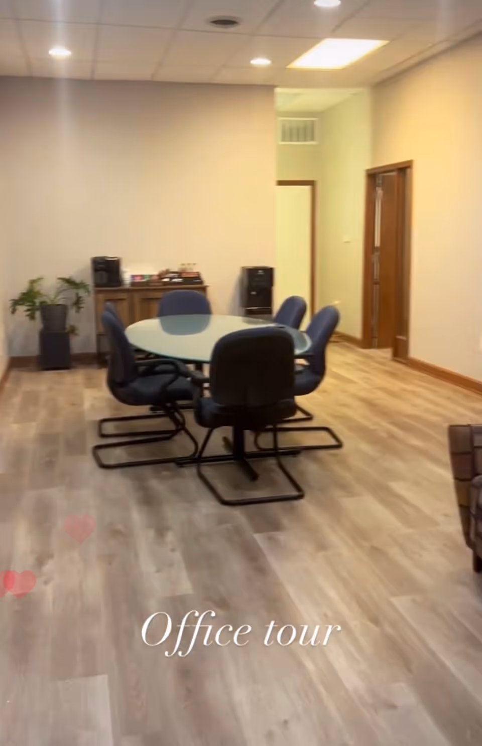 Office-style meeting area with glass round table, blue chairs, plants, and wood flooring.