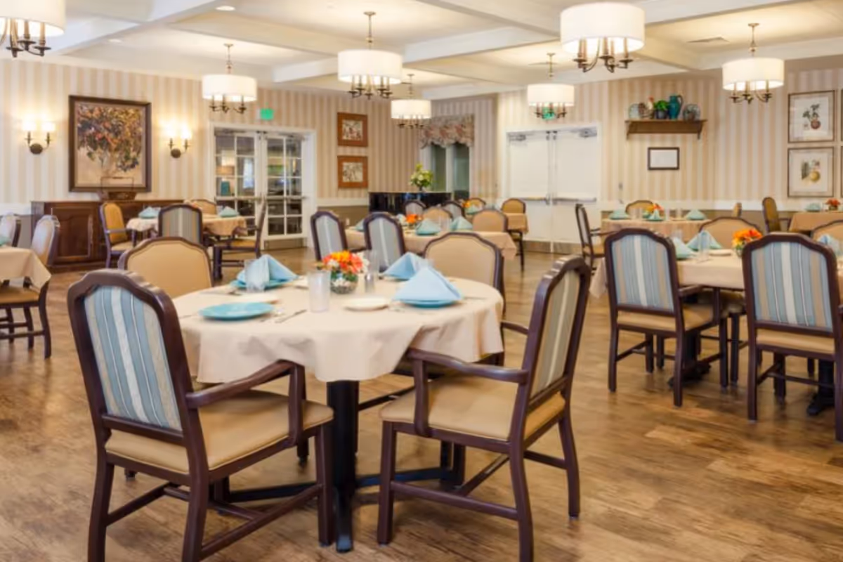 Elegant dining room with round tables, turquoise napkins, and cushioned chairs in a senior living facility.