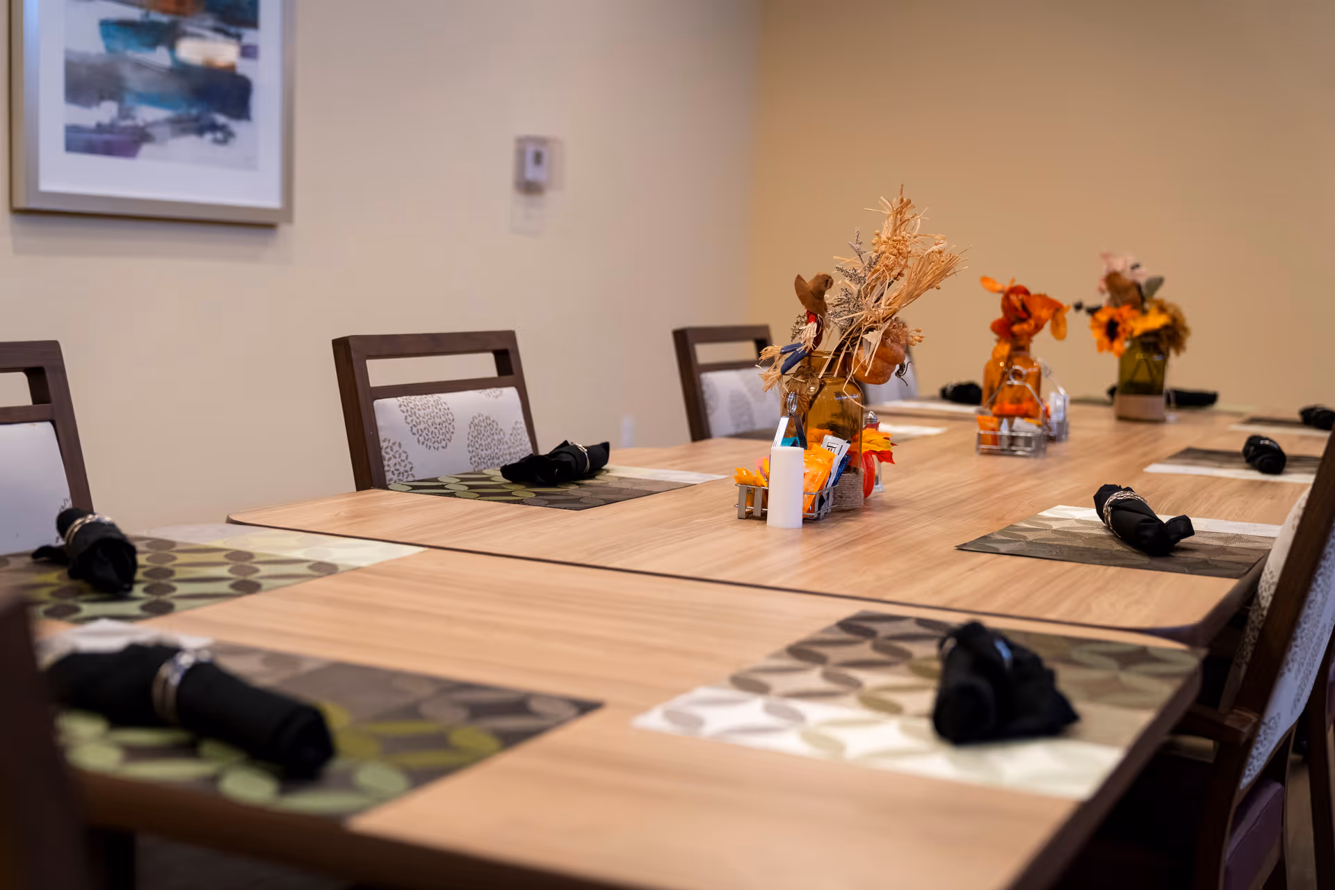 Long dining table set for meals with floral centerpieces in a senior living facility dining room.
