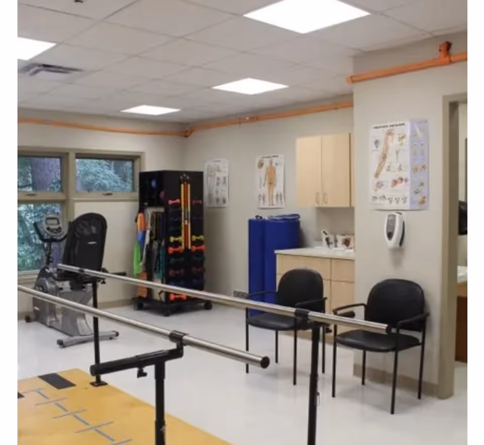 Therapy/exercise room with parallel walking bars, an exercise bike, a rack of weights and bands, chairs, cabinets, and anatomical posters on the walls.