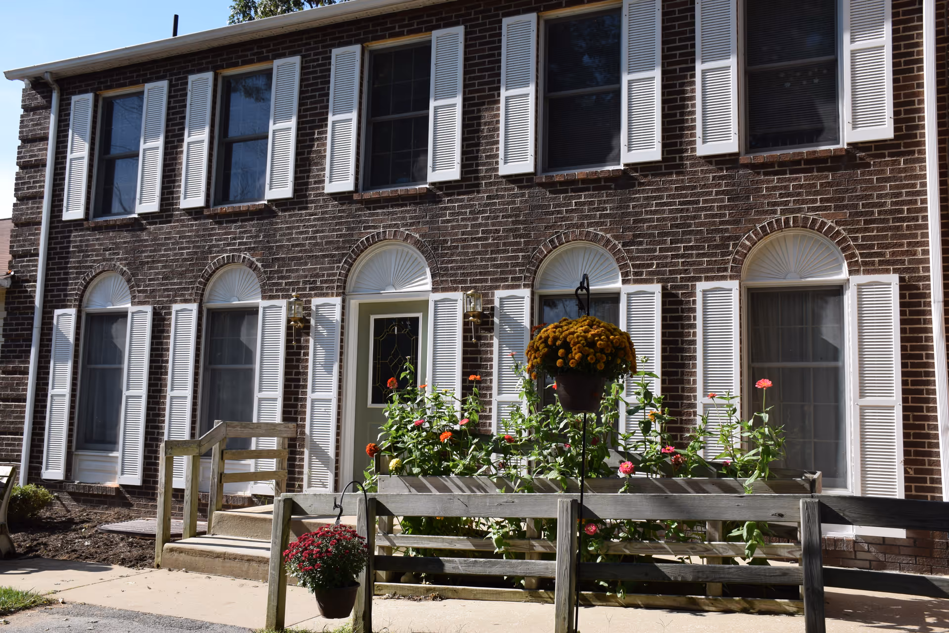 Brick two-story building front with white shutters, a central door, wooden railings and flowering planters at the entrance.