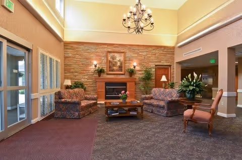 A cozy living room area with a stone fireplace in the center, flanked by two wall sconces and a framed painting above. There are two patterned armchairs and a matching loveseat arranged around a wooden coffee table with flowers and magazines. A wooden chair with a cushion is placed to the right, and there are plants on either side of the fireplace. The room has high ceilings with a decorative chandelier and large windows letting in natural light.