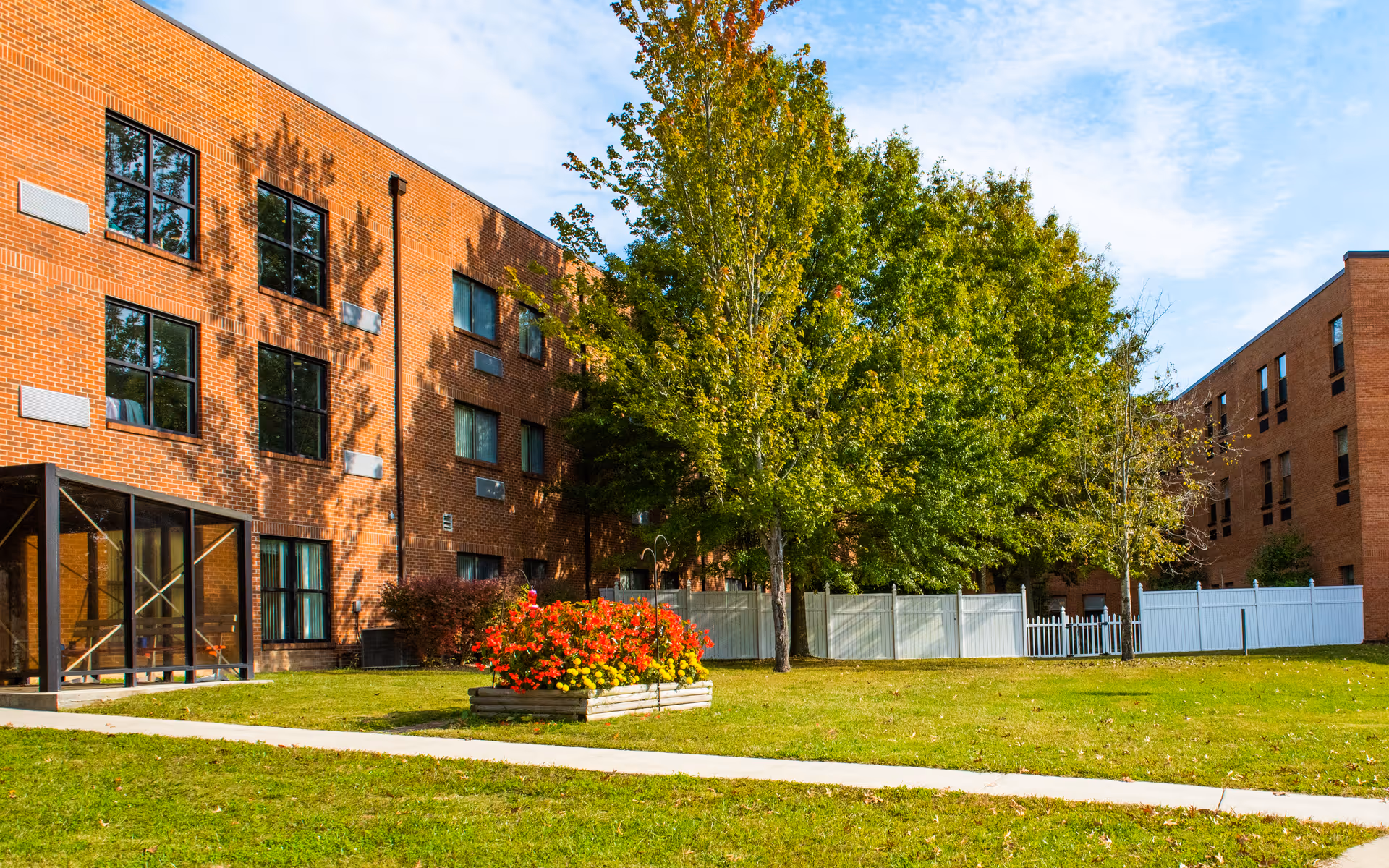 Outdoor view of a senior living facility with red brick buildings, green grass, a flower bed with red and yellow flowers, several trees, and a white fence under a partly cloudy sky.