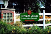 Heritage Village Retirement Campus sign beside a white fence in front of a brick building.