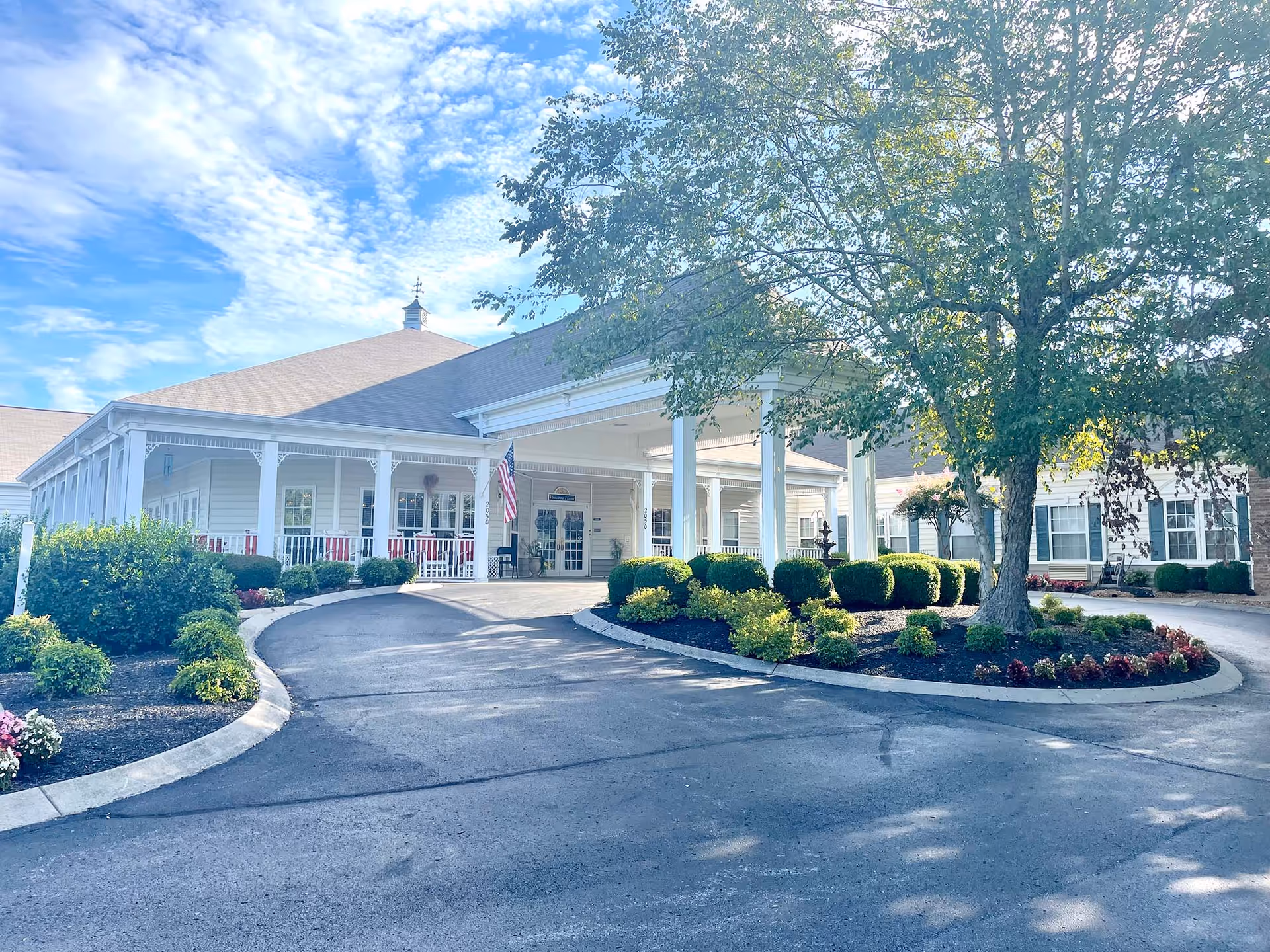 Front exterior view of Morning Pointe of Columbia, a single-story building with a covered entrance, white columns, and a wraparound porch. The driveway curves around landscaped areas with bushes, flowers, and a large tree. The sky is partly cloudy with blue patches.