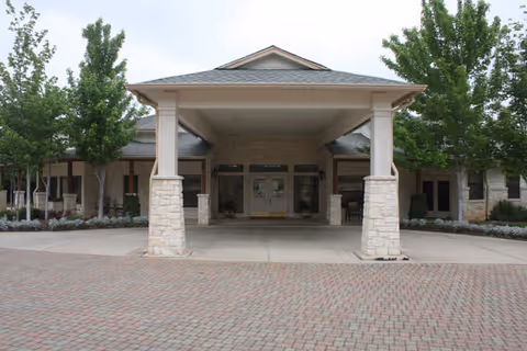 Front exterior view of a single-story building with a covered entrance supported by stone pillars, surrounded by trees and landscaping.