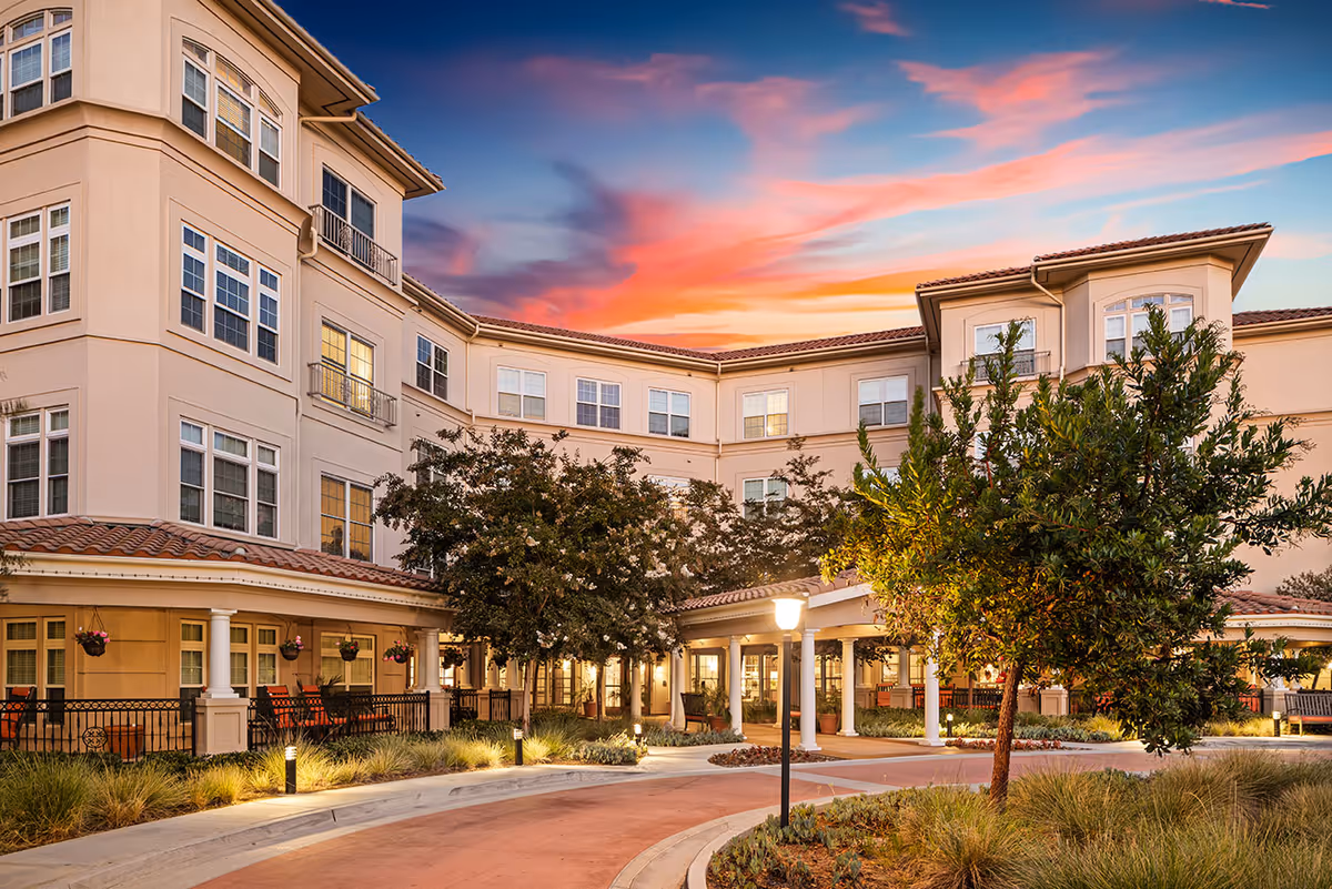 Exterior view of a multi-story senior living facility building at sunset with a curved driveway, landscaped greenery, trees, and outdoor lighting.