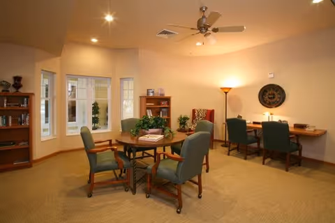 A cozy interior room with a round wooden table surrounded by four green upholstered chairs on casters. There are two bookshelves with books and decorative items, a tall floor lamp, and a wall-mounted clock. The room has beige walls, carpeted floor, recessed ceiling lights, a ceiling fan, and three windows letting in natural light.