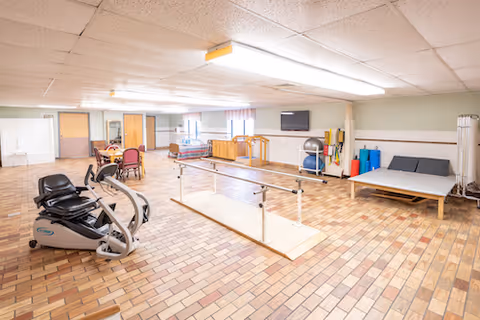 A spacious rehabilitation room with parallel walking bars, exercise machines, therapy tables, and seating.