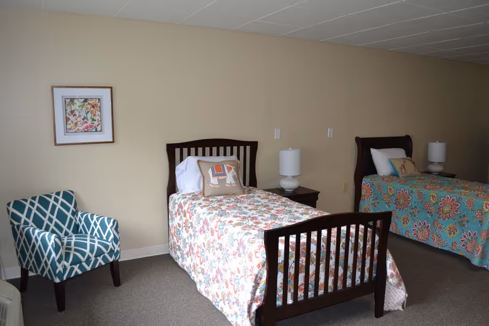 Two single beds with floral bedspreads, nightstands, lamps, and a patterned armchair in a senior living bedroom.
