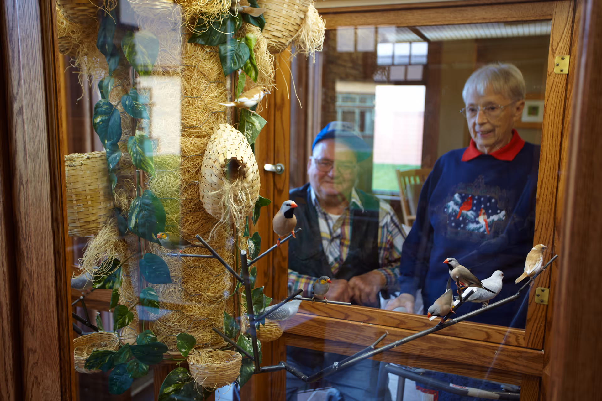 Two elderly residents look through a window at hanging bird decorations inside a senior living center.