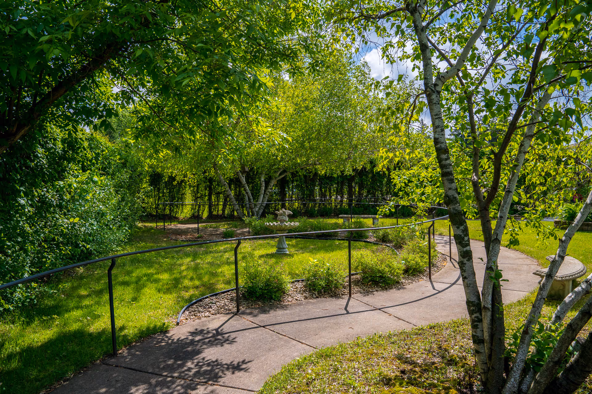 A sunny outdoor garden area with a curved concrete pathway bordered by a black metal handrail. The garden features green grass, bushes, and several trees providing shade. In the center of the garden is a decorative birdbath, and there are stone benches along the pathway.