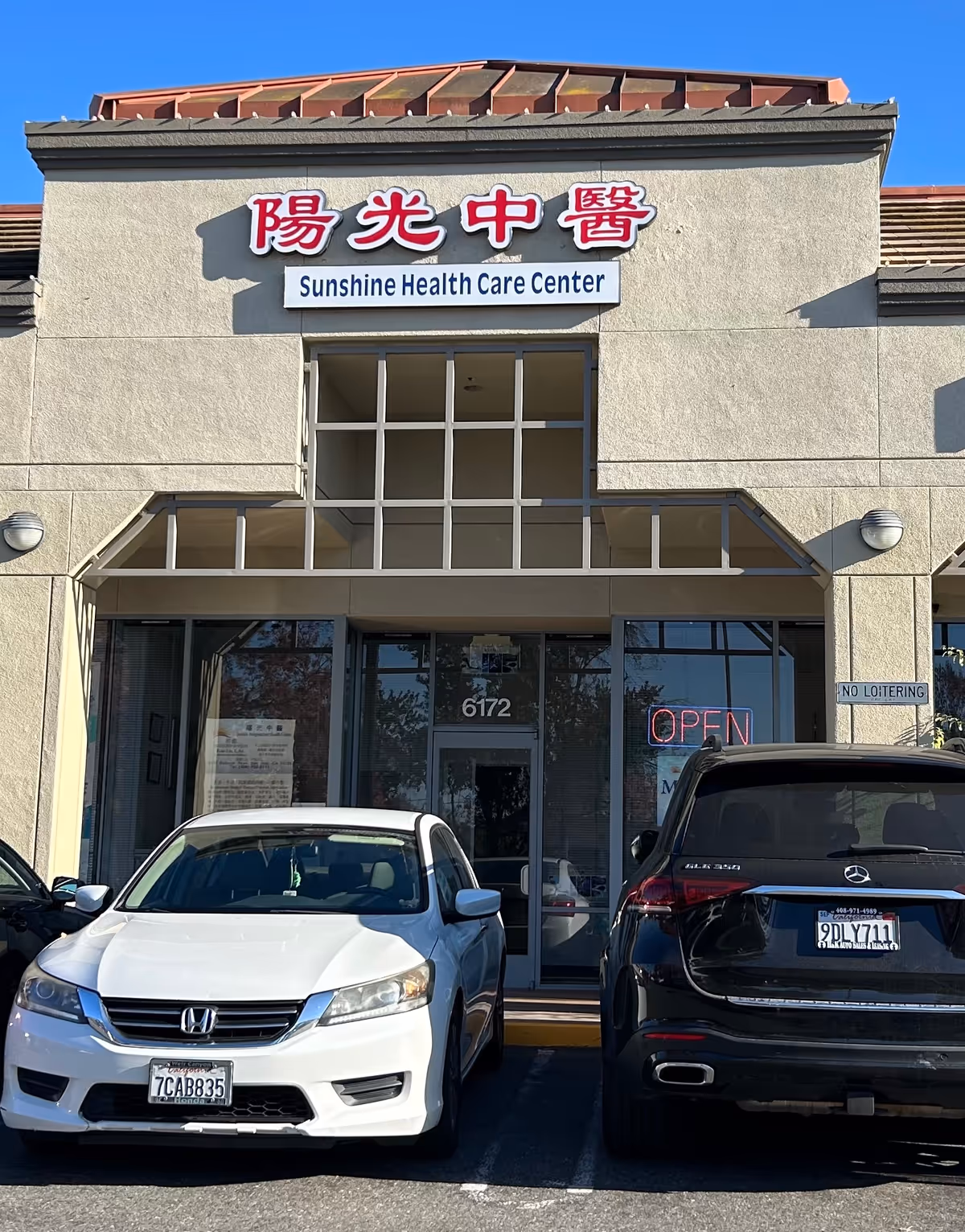 Facade of Sunshine Health Care Center with glass entrance, sign, and two parked cars.