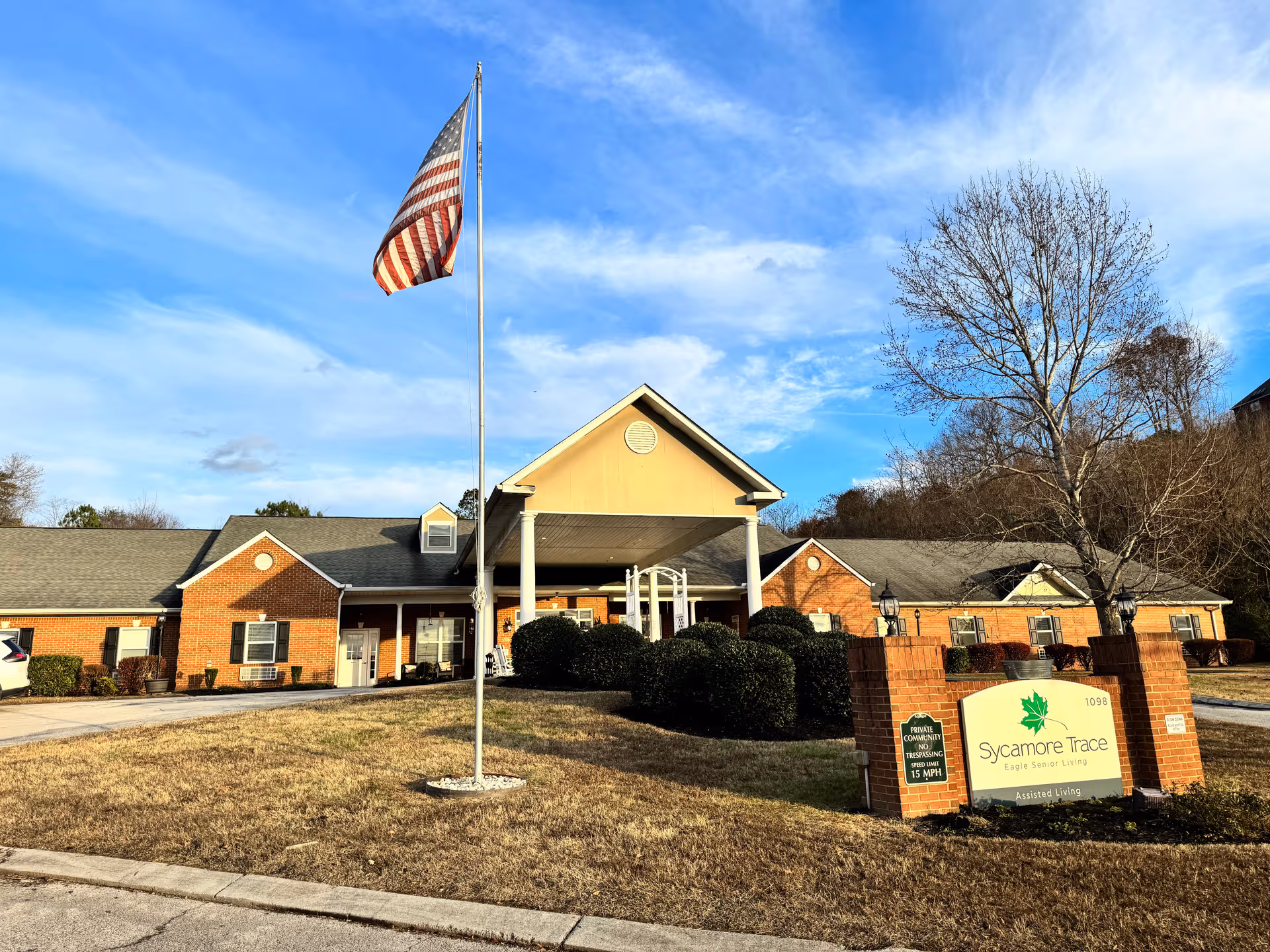 Brick senior living facility with entrance canopy, flagpole, shrubs, and Sycamore Trace sign.