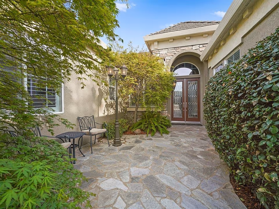 Stone-paved front courtyard with seating, a lamp post, lush shrubs and trees, and double glass entry doors.