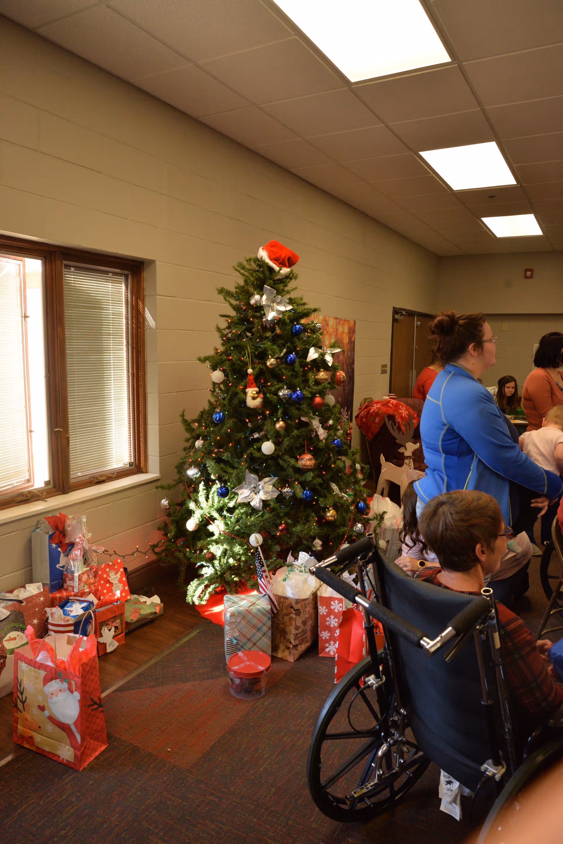 Indoor common room with Christmas tree, gifts, and residents beside a wheelchair.
