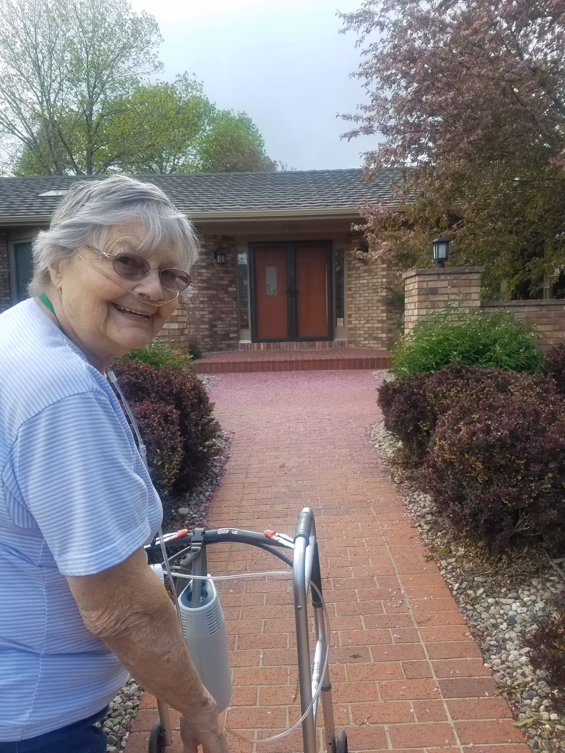 An elderly woman with glasses and a nasal oxygen tube is smiling while using a walker on a brick pathway leading to the front door of a brick house. The pathway is bordered by bushes and small rocks, with trees and greenery in the background.