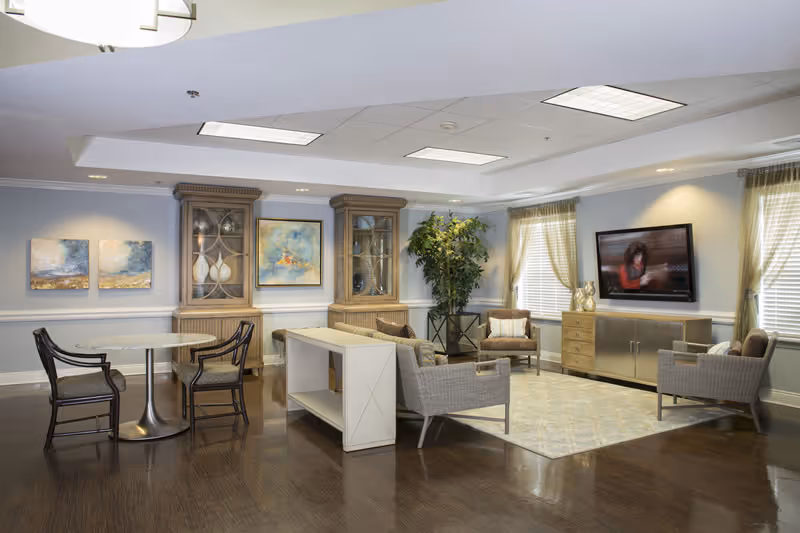 Bright senior living lounge with couches, tables, plants, and wall-mounted television.
