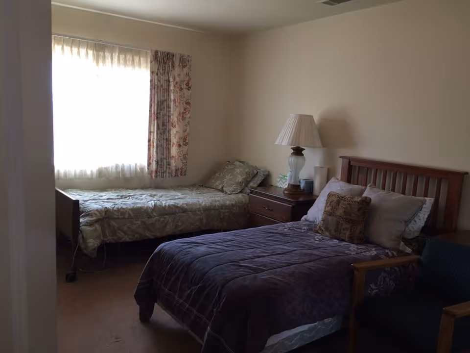 Two-bed bedroom with a wooden headboard, nightstand and lamp beside a window with floral curtains.