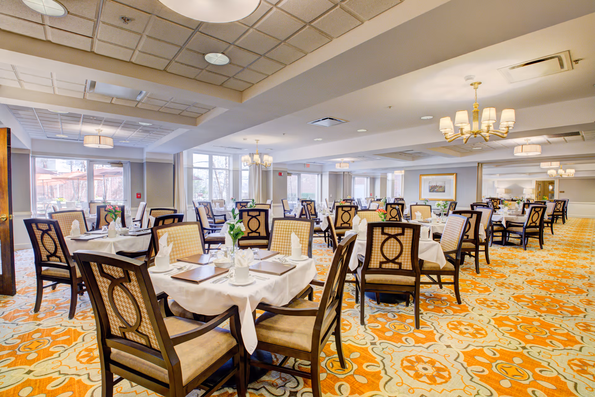 A spacious dining room with multiple round tables covered with white tablecloths, each set with napkins, plates, and menus. The room features patterned orange and beige carpet, large windows letting in natural light, and elegant chandeliers hanging from the ceiling.