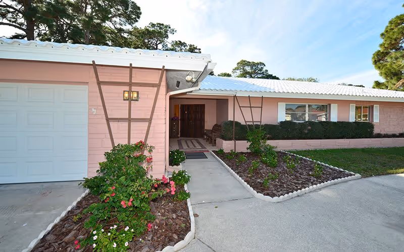 Pink single-story senior living facility with a driveway, garden beds, and a walkway to the entrance.