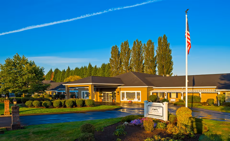 Front exterior of Canterbury Gardens Memory Care Community building with landscaped grounds, entrance sign, and an American flag.