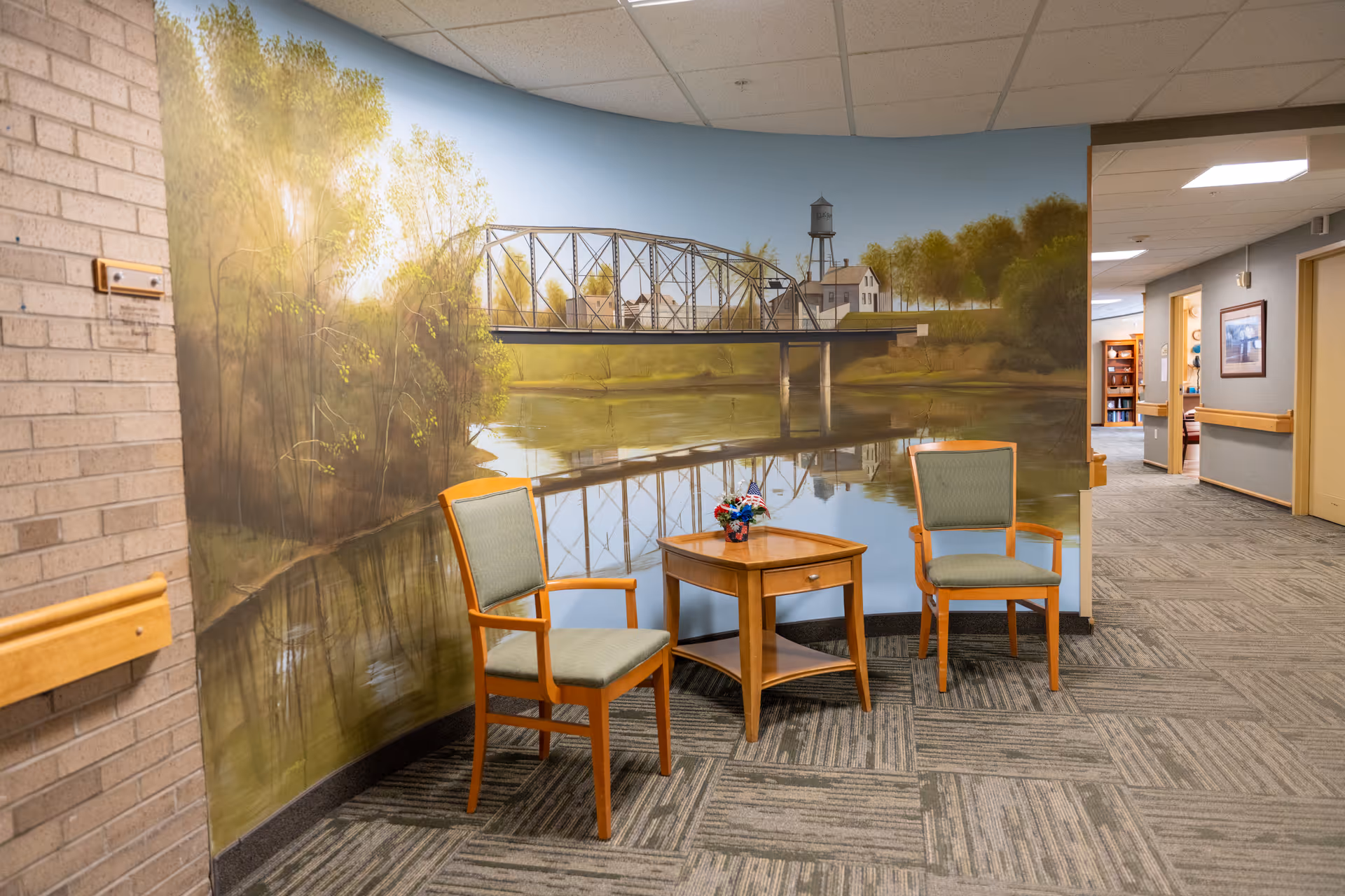 Two wooden chairs and a small table sit before a river mural in a hallway.