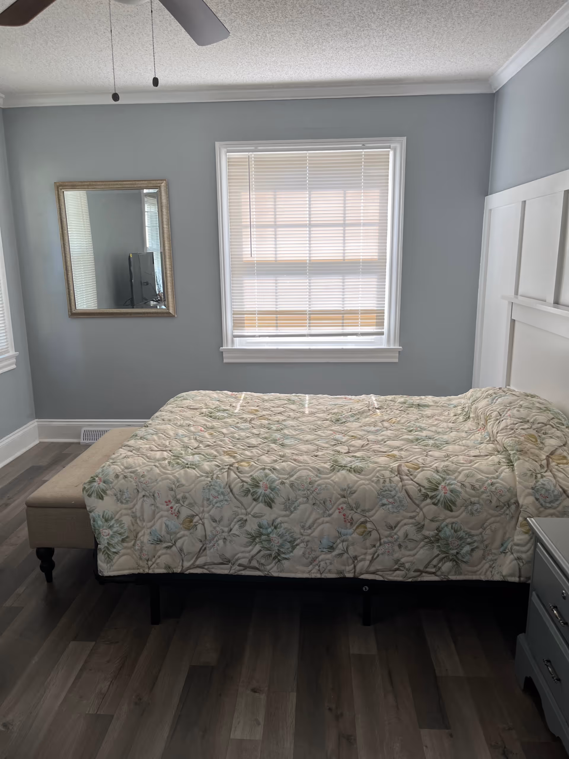A tidy bedroom with a floral quilted bed, window blinds, and a wall mirror.