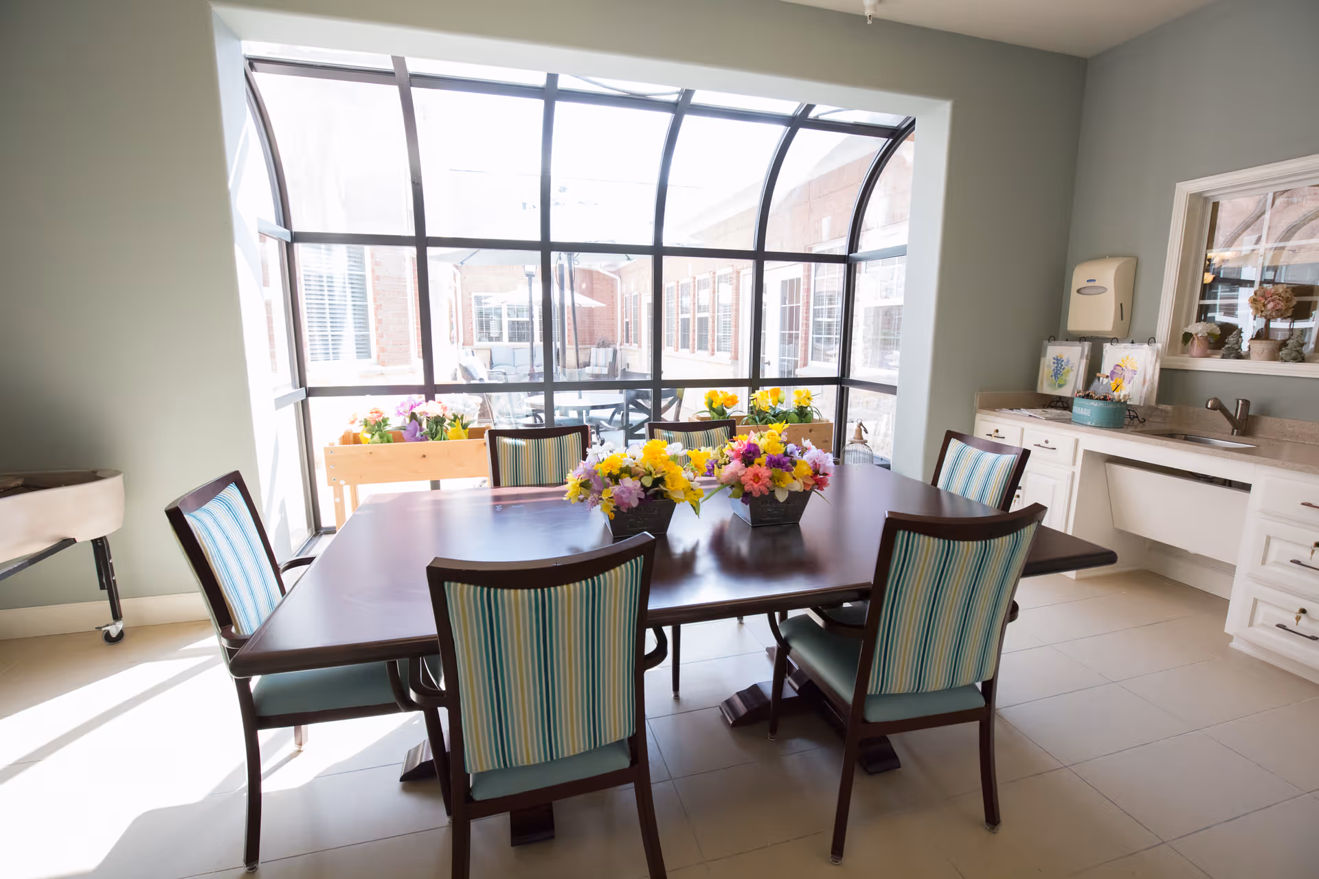 A bright dining area with a dark wooden table surrounded by six chairs with striped upholstery. The table is decorated with colorful flower arrangements. Large curved windows provide a view of an outdoor patio area with additional seating. The room has light-colored tiled flooring and a countertop with a sink and cabinets on one side.