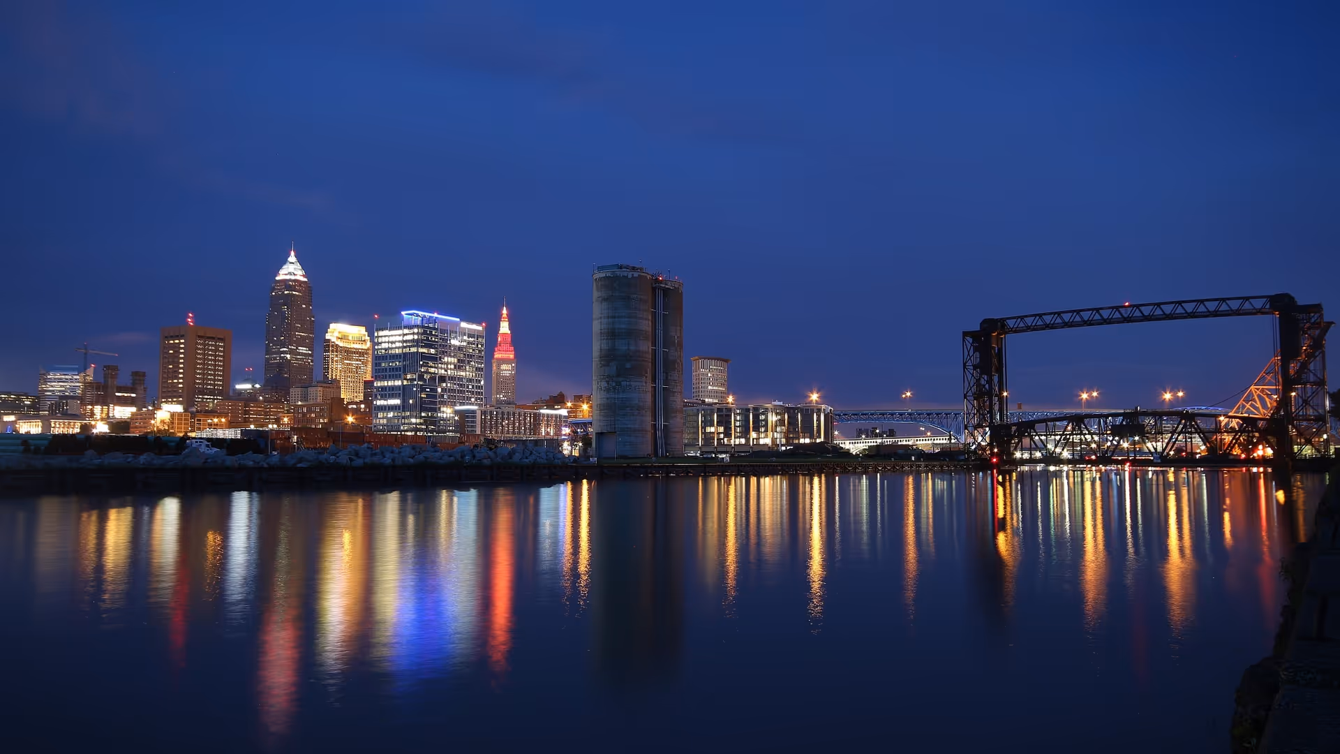 Nighttime city skyline along a calm river, with glowing buildings and a lit bridge.