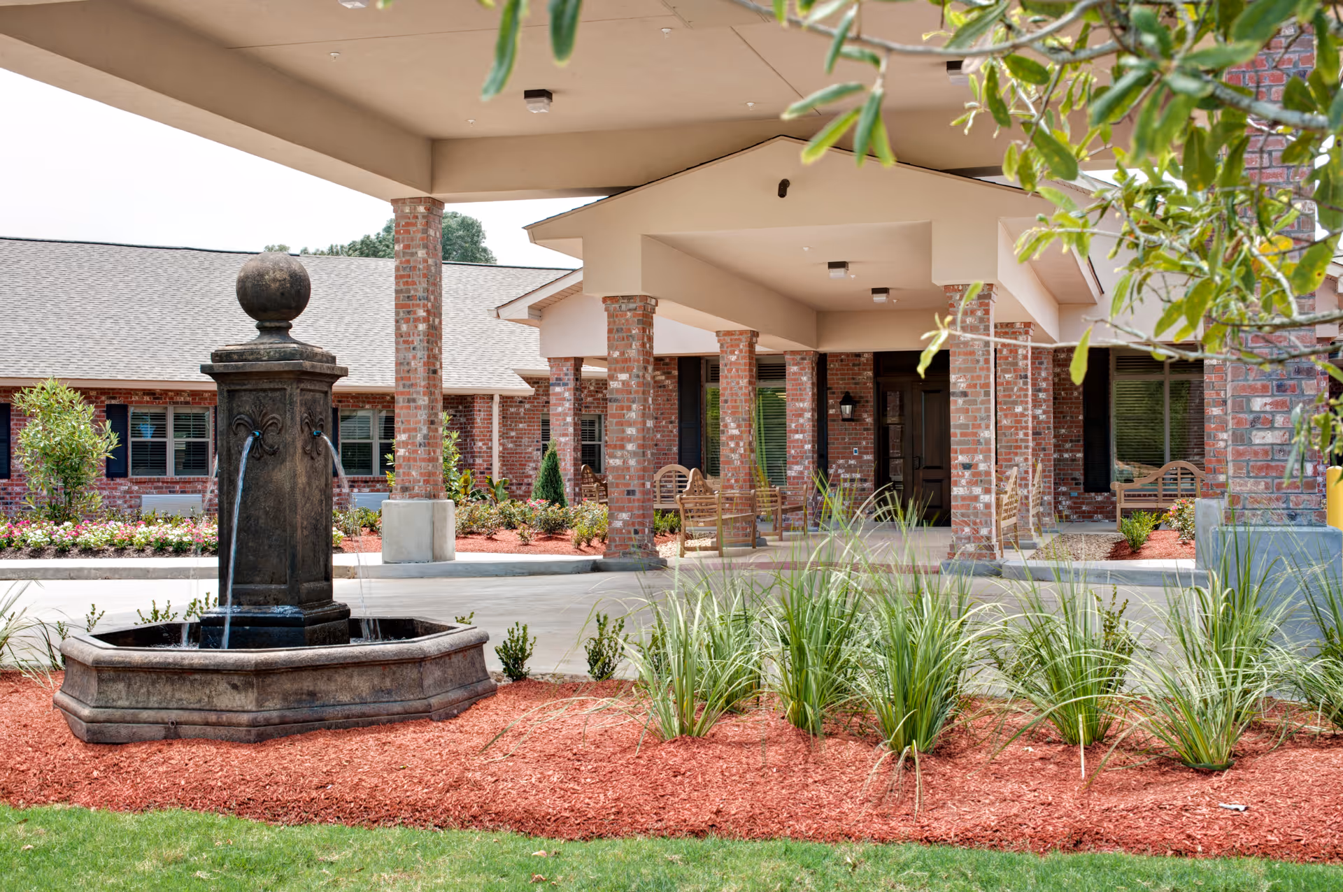 Covered entrance area of a brick building with a decorative water fountain in the foreground, surrounded by landscaping with green plants and red mulch. Wooden benches are placed under the covered area near the entrance.