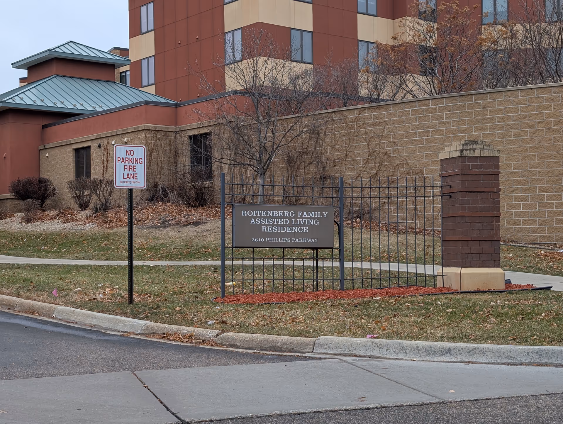Front exterior of the Roitenberg Family Assisted Living residence with a metal sign and a 'No Parking Fire Lane' sign on the lawn.