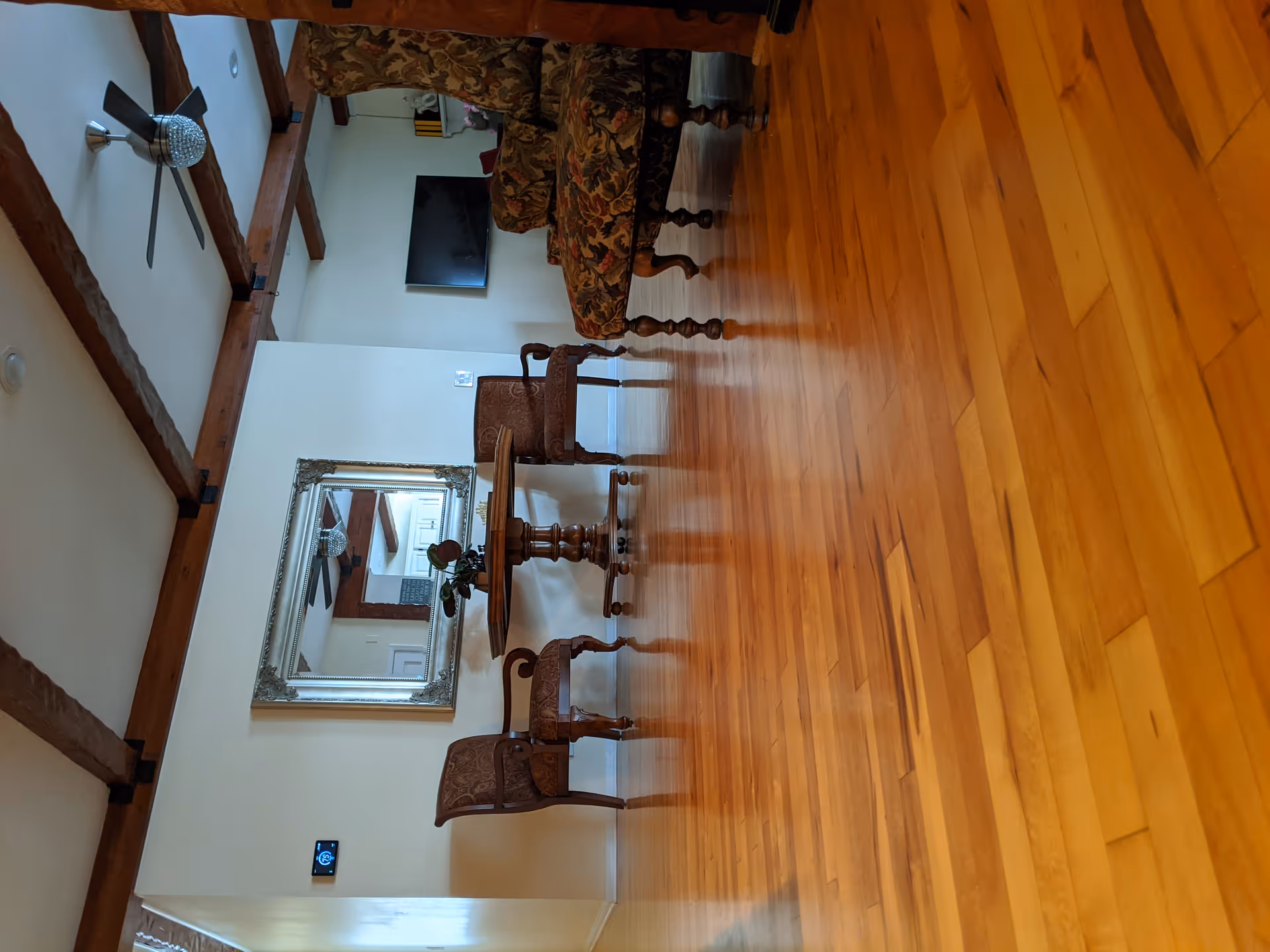 Interior living room with polished hardwood floors, a round wooden table with ornate chairs, a sofa, wall mirror, TV, and exposed ceiling beams.