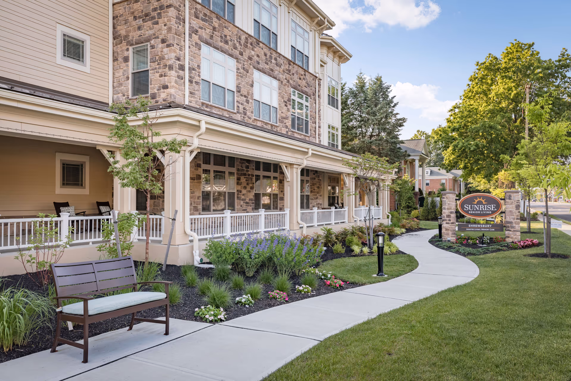 Exterior of Sunrise of Shrewsbury senior living with a curved sidewalk, porch seating, and landscaped garden.