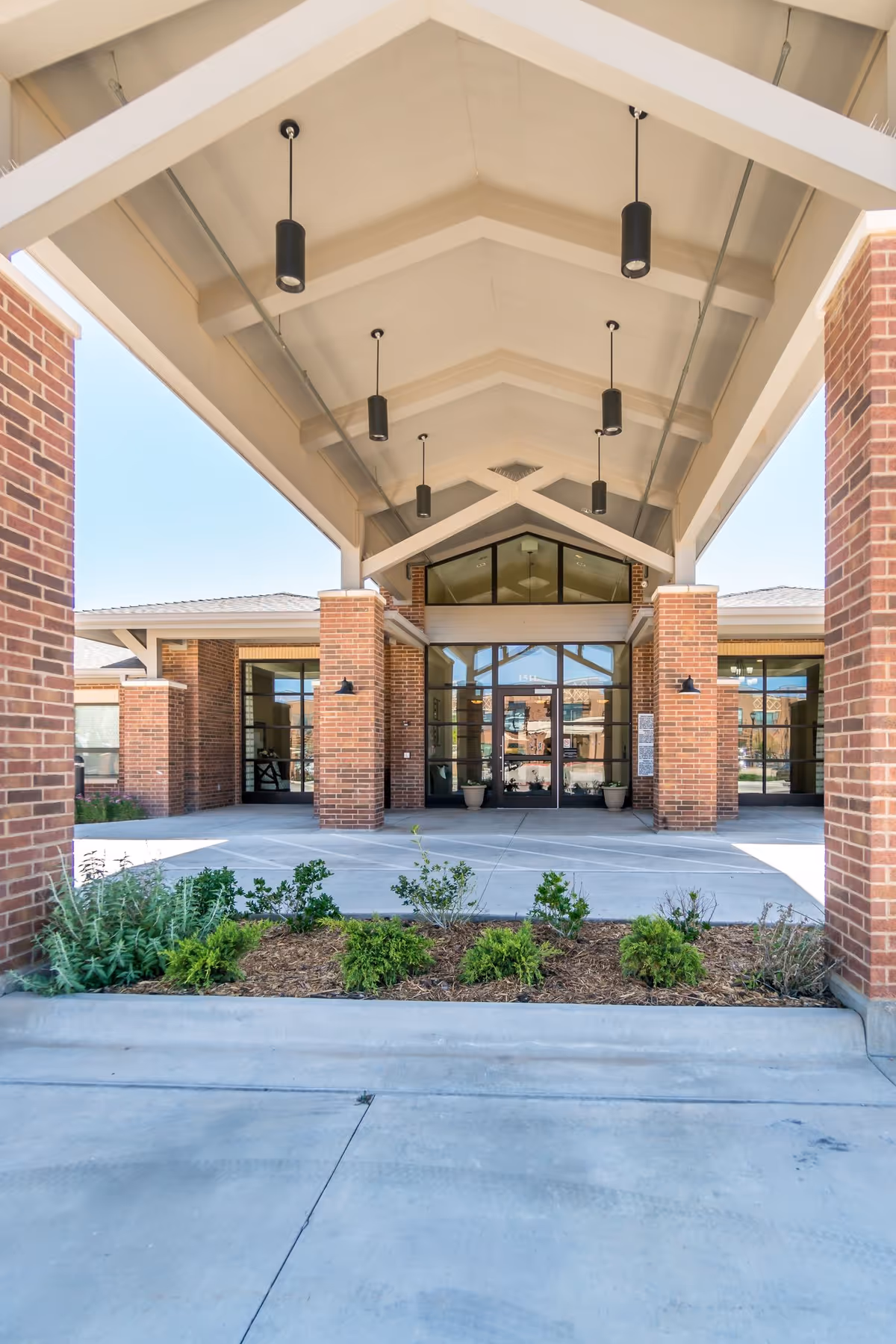 Entrance of a building with a covered walkway supported by brick pillars and a high ceiling with hanging lights. There are glass doors at the entrance and small plants in a landscaped area in front.