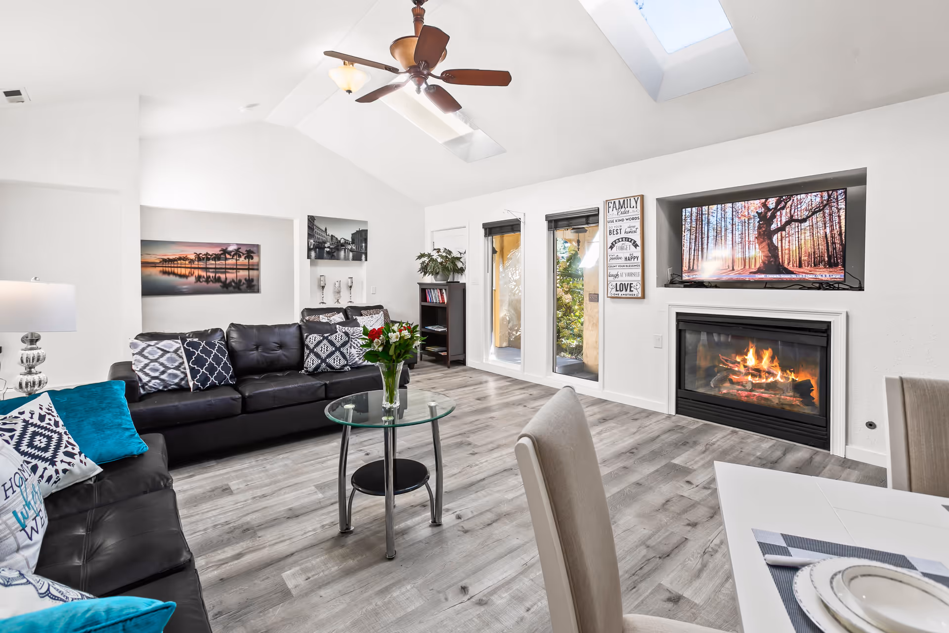Bright modern living room with black leather sofas, a glass coffee table, skylights, a fireplace with TV above, and a small dining area.