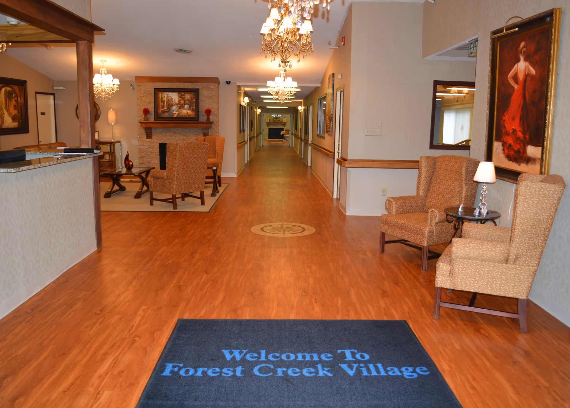 Interior lobby hallway of Forest Creek Village with hardwood floors, seating areas, chandeliers, and a welcome mat.