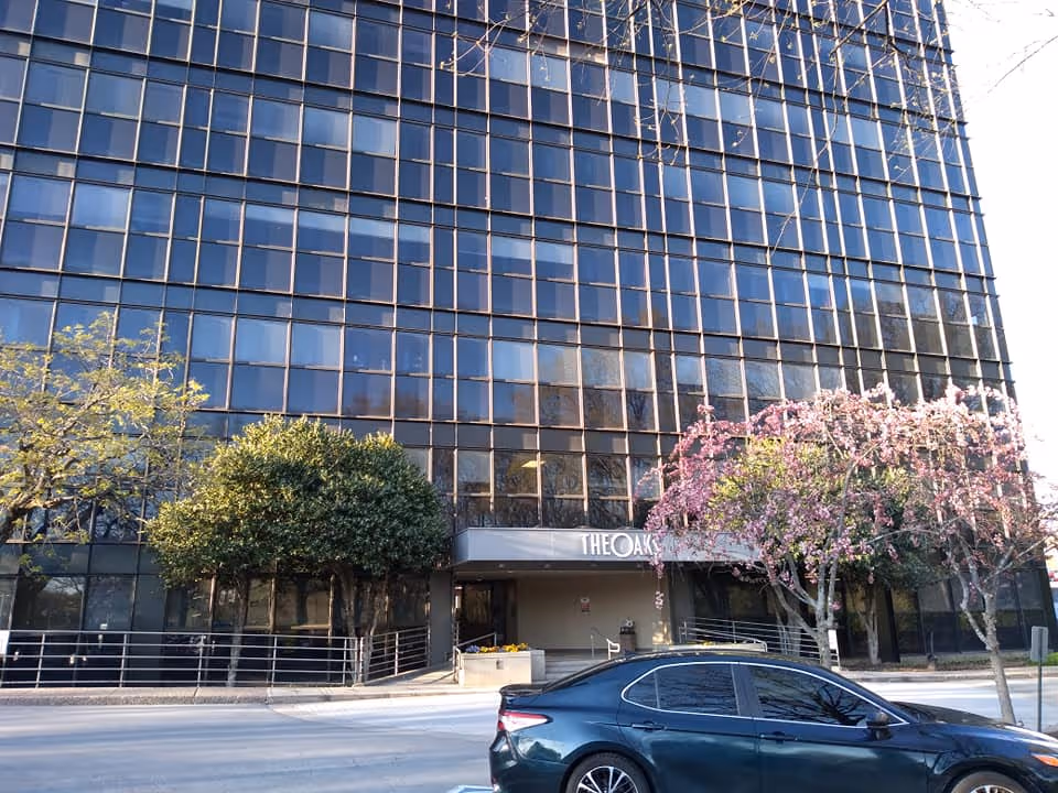 Tall glass-front building facade with entrance, trees, and a parked car in front.
